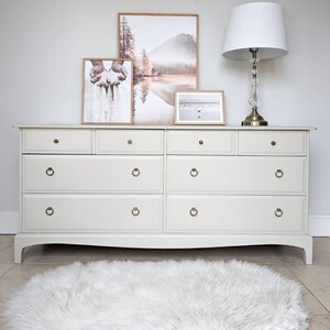 May include: A cream-coloured chest of drawers with multiple drawers and brass handles. Three framed landscape prints and a lamp with a white shade sit on top. A fluffy white rug is in front.