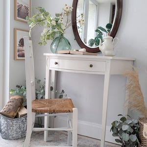 May include: A white dressing table with a drawer, oval mirror, and a teapot. A white chair with a woven seat is in front of the table. The room is decorated with plants, framed pictures, and a decorative bucket.