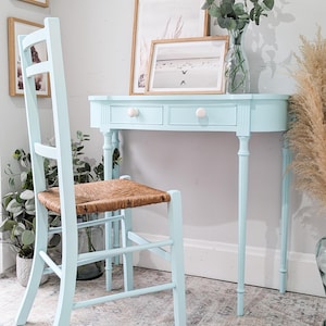 May include: A pale blue desk with two drawers and a matching chair with a woven seat. The desk has a curved front and is paired with a tall-backed chair. Framed artwork and a vase of greenery complete the scene.