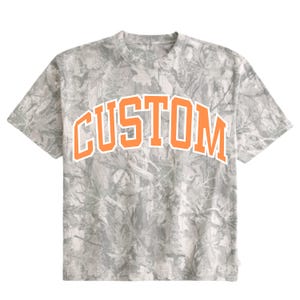 Könnte beinhalten: Ein kurzärmeliges T-Shirt mit einem grau-weißen Tarnmuster aus Blättern und Zweigen. Das Wort CUSTOM ist in einer großen, fetten, orangefarbenen Schrift im College-Stil mit weißer Umrandung mittig auf der Brust des T-Shirts aufgedruckt.