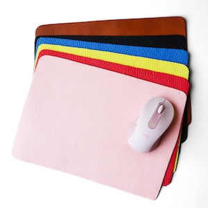 May include: A stack of colorful leather mouse pads in brown, black, blue, yellow, red, and pink. A white wireless mouse with a pink scroll wheel is on top of the pink mouse pad.