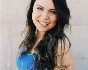 May include: A woman with long dark hair and blue eyes smiles at the camera. She is wearing a blue tank top with the words "Mermaid" written in gold glitter.