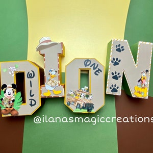 May include: Wooden letters spelling "DION" decorated with a jungle theme. The letter "D" has a Mickey Mouse character, the letter "I" has Donald Duck, the letter "O" has a safari jeep with animal characters, and the letter "N" has Goofy. The letters are decorated with paw prints and the word "WILD" is written on the letter "D". The letters are on a green and brown background.