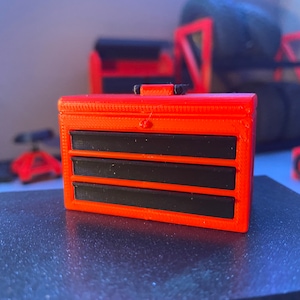 May include: A red metal toolbox with three drawers. The drawers have black fronts and are closed. The toolbox is on a dark gray surface.