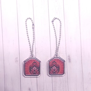 May include: Two clear acrylic keychains with a red and black design. Each keychain has a silver ball chain and a geometric shape with a decorative pattern. The background is a light wood surface.
