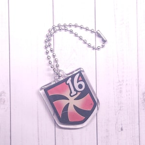May include: A clear acrylic keychain with a shield design. The shield features a red and white burst pattern with the number 16 in white. It hangs from a silver ball chain.