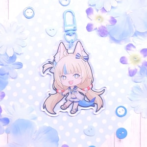 May include: A clear acrylic charm featuring a cartoon character with fox-like ears, a blue tail, and a sailor-style outfit. The charm is attached to a light blue clasp. The background includes blue flowers, buttons, and a polka-dot pattern.