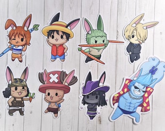 May include: A collection of eight cartoon stickers featuring characters from a popular anime series, each reimagined as a bunny. The stickers showcase various characters with unique outfits and accessories, including carrots and hats. The stickers are on a white wooden background.
