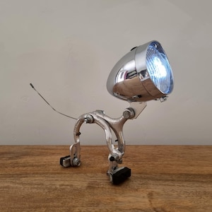 Pode incluir: Um candeeiro de mesa construído a partir de um conjunto de pinças de travão de bicicleta em tom prateado e uma caixa de farol cromada em forma de bala. O candeeiro apresenta uma lente brilhante iluminada e um fio fino e escuro na parte traseira.