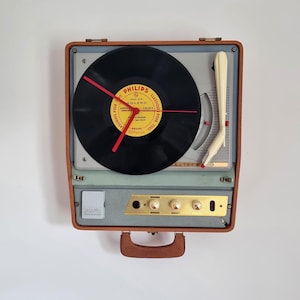 Op de afbeelding: Een wandklok gemaakt van een vintage draagbare platenspeler. De wijzerplaat is een zwarte vinylplaat met een geel Philips Bolero-label en drie rode wijzers. De speler heeft een bruine behuizing, een crèmekleurige toonarm en een goudkleurig bedieningspaneel.