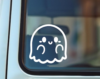 May include: A white vinyl decal of a cartoon ghost with a smiling face and wavy bottom edge is affixed to the rear window of a silver vehicle. The ghost has two large eyes and a small mouth. The background is a blurred street scene.