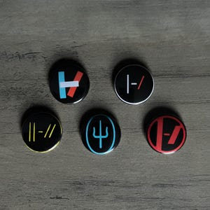 May include: Five black round buttons with various graphic designs in red, white, blue, and yellow. The buttons are arranged on a wooden surface. The designs include geometric shapes and symbols.