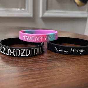 May include: Three silicone wristbands. One is a pink, blue, and purple gradient with the white text TWENTY ØNE. Another is black with white pixelated text. The third is black with the white handwritten-style text Push on through.