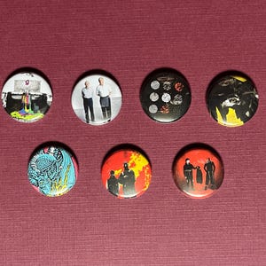 May include: A collection of seven round pinback buttons with various designs. The buttons feature images of people, abstract art, and graphic designs in a variety of colors including black, red, yellow, and blue. The buttons are arranged on a maroon surface.