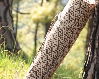 May include: Rolled yoga mat in a patterned carrying case. The case is beige with a repeating brown geometric design. A matching strap is attached for easy carrying. The mat is leaning against a tree in a natural outdoor setting.