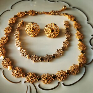May include: A gold-tone necklace and earrings set featuring a delicate filigree design. The necklace is a chain with a repeating pattern of small squares and swirls. The earrings are round with a similar swirl design.