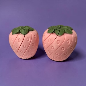 May include: Two strawberry-shaped objects with light pink bodies featuring diagonal grooves and oval indentations. Each piece is topped with a dark green, multi-petaled stem component. The items have a smooth, matte finish and a segmented appearance.