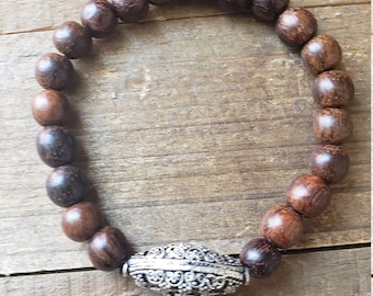 May include: A brown wooden beaded bracelet with a silver charm. The charm has a detailed floral design.