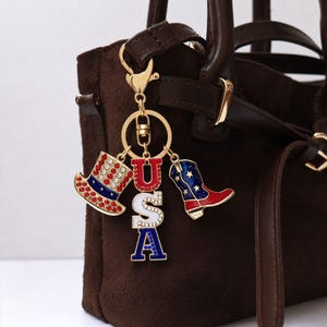 Op de afbeelding: Een goudkleurige sleutelhanger met patriottische bedels. Bedels zijn onder meer een rood-wit-blauwe hoge hoed, een rood-wit-blauwe cowboylaars en de letters "USA". De sleutelhanger is bevestigd aan een bruine handtas.
