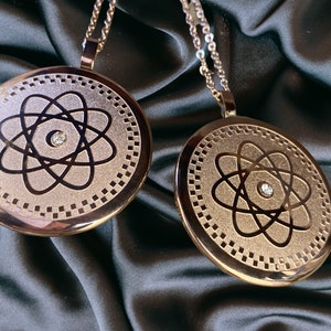 May include: Two gold-toned pendants with a silver atom design and a small clear rhinestone in the center. The pendants are on a black satin background.