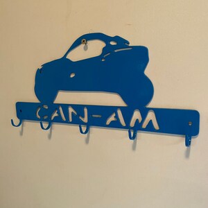 May include: A blue metal key holder in the shape of a Can-Am vehicle. The key holder has the words "CAN-AM" cut out and includes four hooks for keys. It is mounted on a white wall.