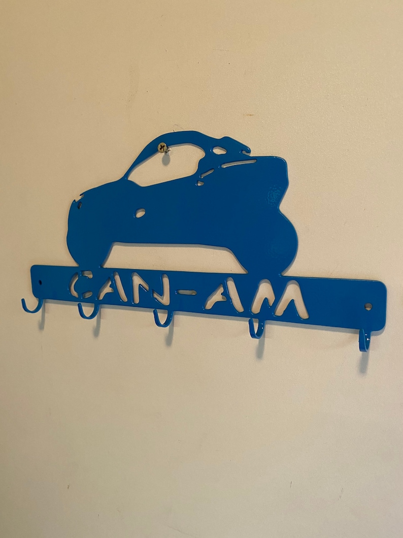 May include: A blue metal key holder in the shape of a Can-Am vehicle. The key holder has the words "CAN-AM" cut out and includes four hooks for keys. It is mounted on a white wall.