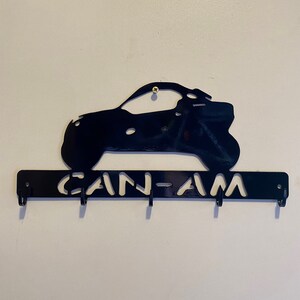 May include: Black metal key holder with a cut-out design of an off-road vehicle and the text "CAN-AM". The key holder has four hooks for keys and is mounted on a white wall.