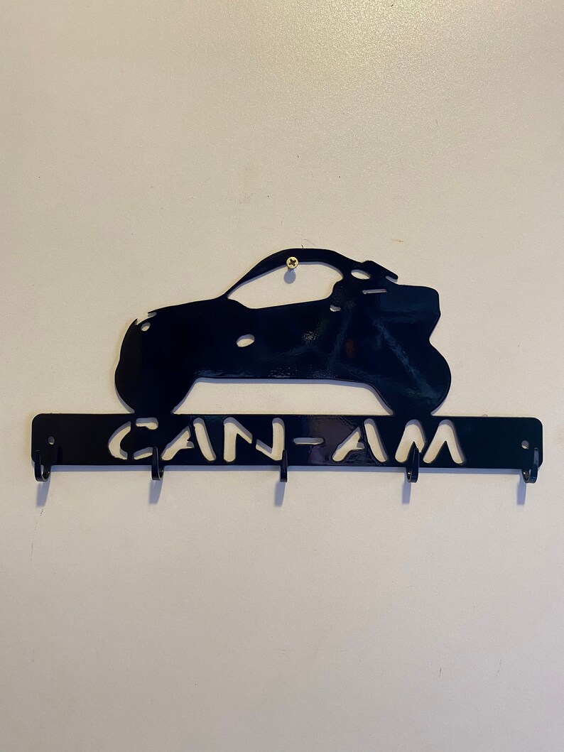 May include: Black metal key holder with a cut-out design of an off-road vehicle and the text "CAN-AM". The key holder has four hooks for keys and is mounted on a white wall.