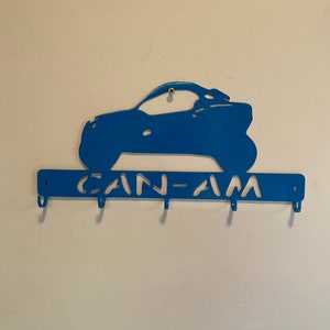 May include: A blue metal key holder in the shape of an off-road vehicle. The key holder has the text "CAN-AM" and four hooks for keys. It is mounted on a white wall.