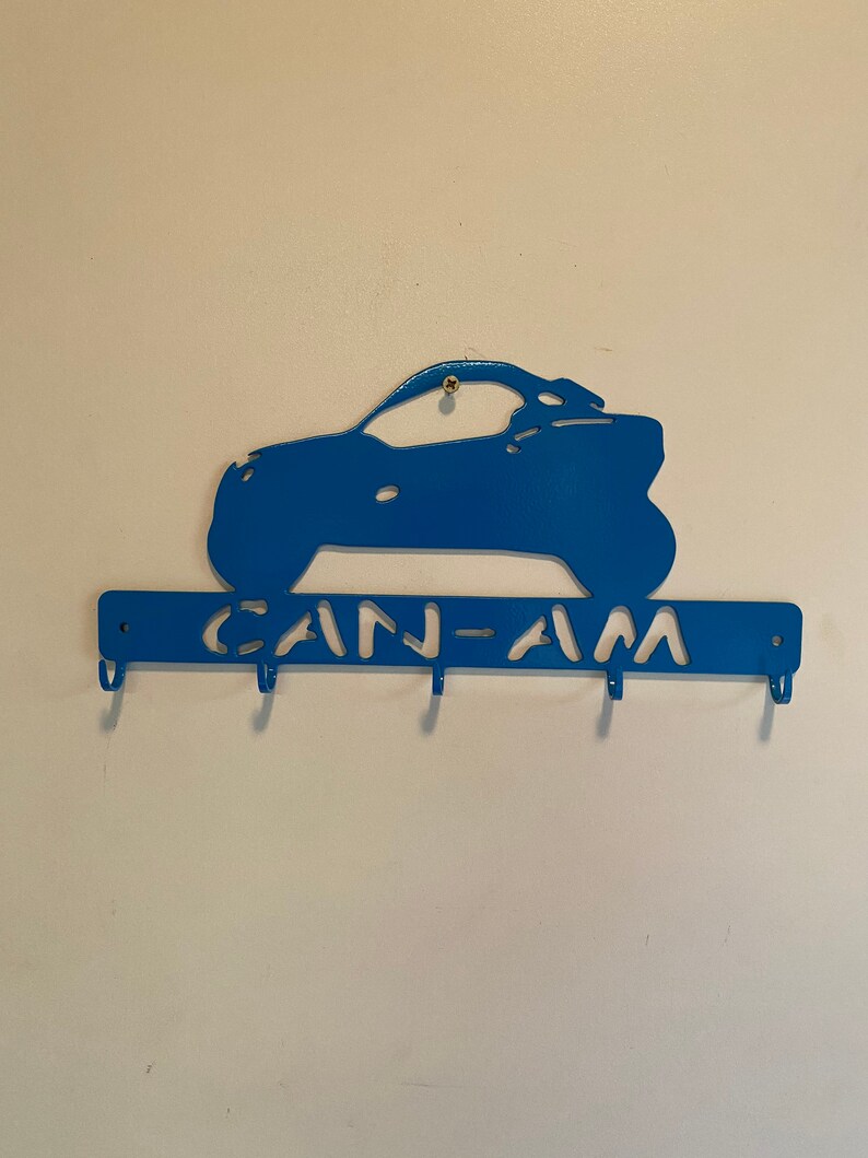 May include: A blue metal key holder in the shape of an off-road vehicle. The key holder has the text "CAN-AM" and four hooks for keys. It is mounted on a white wall.