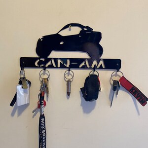 May include: Black metal key holder with a vehicle silhouette and the cut-out text "CAN-AM". Various keys and keyrings are suspended from the hooks. The key holder is fixed to a white wall.