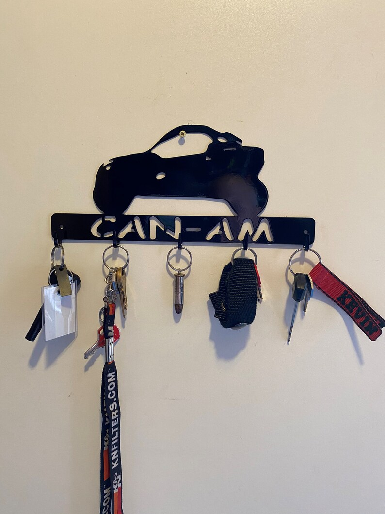 May include: Black metal key holder with a vehicle silhouette and the cut-out text "CAN-AM". Various keys and keyrings are suspended from the hooks. The key holder is fixed to a white wall.