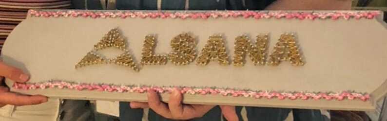 May include: A wooden sign with a pink and white border, the name "ALSANA" is spelled out in gold tacks.