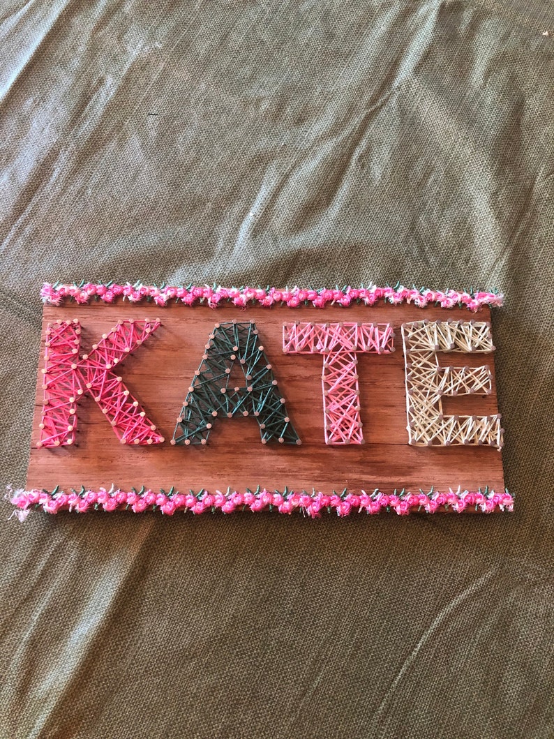 May include: A wooden sign with the name "KATE" spelled out in colorful string art. The letters are surrounded by a pink and green border.