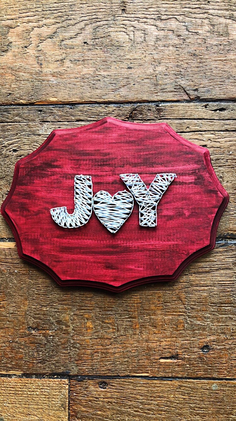 May include: A red wooden sign with the word "JOY" spelled out in white string. The letters are surrounded by a white heart.