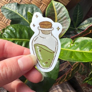 May include: A sticker featuring a cartoon illustration of a potion bottle. The bottle is clear with a cork stopper and contains a green liquid. Small sparkles surround the bottle, suggesting a magical theme. The sticker is held by a person's hand.