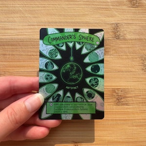 May include: A rectangular trading card titled Commander's Sphere featuring a central green globe icon surrounded by twelve eye-shaped illustrations on a black background. The card includes text detailing its function as an artifact and game rules.