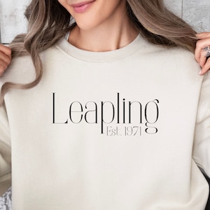 May include: A cream colored sweatshirt with the text "Leapling Est. 1971" printed on the front.