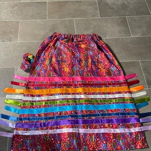 May include: A vibrant, patterned skirt with colorful ribbons. The skirt features a floral design in shades of red, pink, and blue. Horizontal ribbons in various colors, including orange, green, and purple, are attached. A matching scrunchie is also visible.