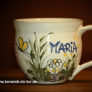Könnte beinhalten: Eine weiße Keramiktasse mit einem Blumendesign und dem Namen "Maria" auf der Seite. Die Tasse hat einen abgerundeten Körper und einen Henkel.
