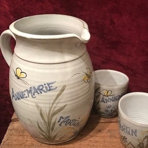 Könnte beinhalten: Eine weiße Keramikkanne mit Henkel und Ausguss, verziert mit einem Schmetterling, Gras und den Namen "Annemarie" und "Martin". Zwei kleinere weiße Keramikbecher mit Schmetterlings- und Namensdekorationen sind ebenfalls abgebildet.