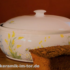 Könnte beinhalten: Weiße Keramik-Brottöpfe mit floralem Design mit gelben Blumen und grünen Blättern. Die Brottopf hat einen Deckel mit einem Knopf oben. Ein Laib dunkelbraunes Brot steht vor dem Brottopf. Der Text "www.keramik-im-tor.de" ist unten im Bild sichtbar.