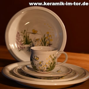 Könnte beinhalten: Ein Keramik-Geschirrset mit einer großen Schüssel, einer Tasse und Tellern. Die weißen Teile sind mit einem floralen Design in Gelb, Grün und Lila verziert. Die Website-Adresse "www.keramik-im-tor.de" ist oben sichtbar.