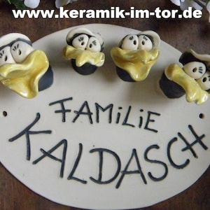 Könnte beinhalten: Ein weißes ovales Schild aus Keramik mit dem Text "Familie Kaldasch" in schwarzer Schrift. Vier kleine Keramikfiguren von Comicvögeln mit gelben Schnäbeln sind an dem Schild befestigt.