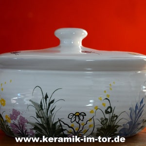 Könnte beinhalten: Weiße Keramik-Servierplatte mit Deckel, verziert mit einem Blumendesign in Grün-, Gelb- und Lilatönen. Die Platte hat zwei Griffe an den Seiten und den Text "www.keramik-im-tor.de" auf der Unterseite.