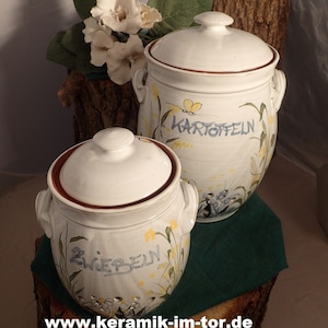 Könnte beinhalten: Zwei weiße Keramik-Behälter mit Deckeln, einer mit der Aufschrift "Zwiebeln" und der andere mit "Kartoffeln". Beide Behälter haben einen braunen Rand und sind mit gelben Blumen und grünen Blättern verziert.