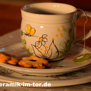 Könnte beinhalten: Eine weiße Keramik-Teetasse mit floralem Design und einem Schmetterling, eine Untertasse und drei Kekse auf einem weißen Teller. Die Teetasse hat eine Schnur an dem Henkel, an der ein Teebeutel hängt. Der Text "www.keramik-im-tor.de" ist am unteren Rand des Bildes sichtbar.