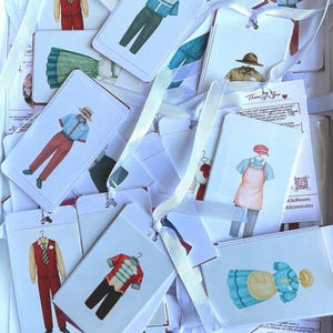 May include: A collection of white rectangular tags with illustrations of various outfits, including uniforms and historical clothing. Each tag is adorned with a white ribbon. The tags feature a variety of colors, including red, blue, and green.