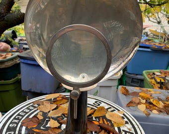 Puede incluir: Una lámpara lupa vintage con un reflector grande, redondo y plateado y una lente de aumento redonda más pequeña. La lámpara tiene una base negra en forma de trompeta.