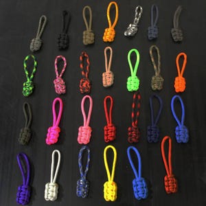 May include: A collection of twenty-eight zipper pulls made from braided paracord in various solid colors and patterns, including neon green, pink, orange, blue, yellow, black, grey, white, and camouflage, each featuring a looped cord attachment.